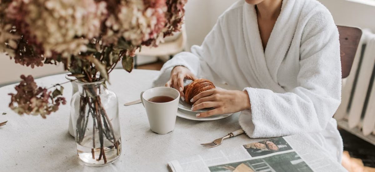 A woman in a bathrobe having breakfast indoors with a croissant and tea.