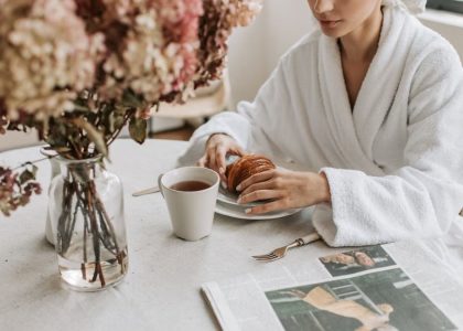 A woman in a bathrobe having breakfast indoors with a croissant and tea.