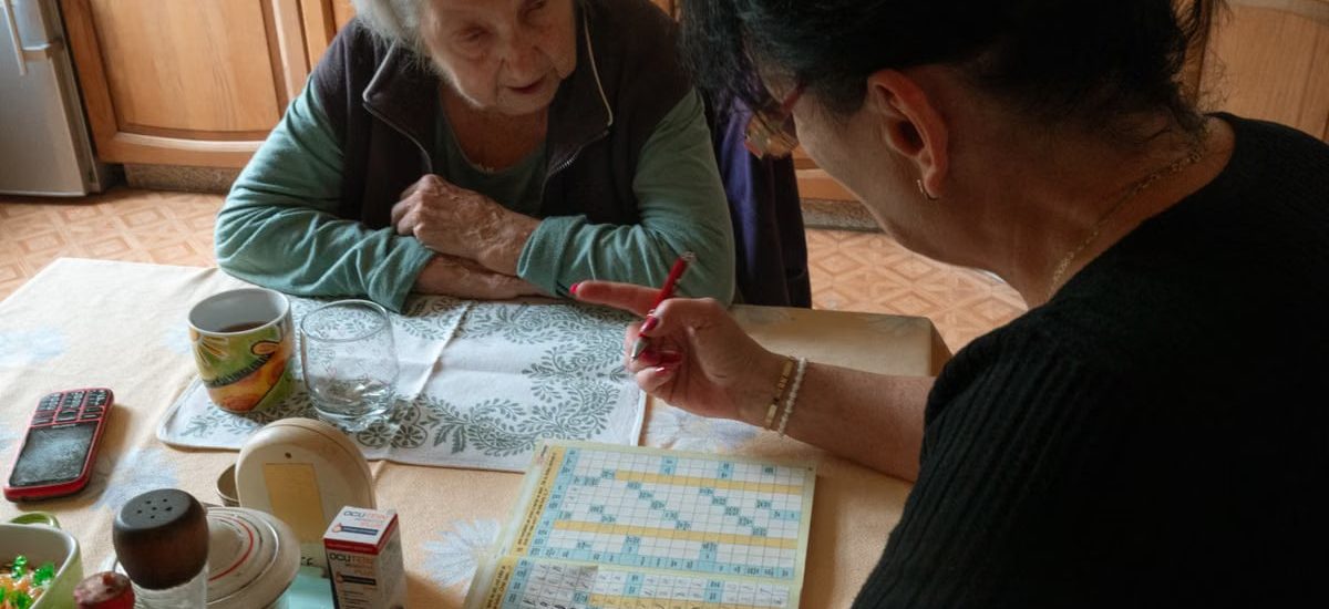 A senior woman and her caregiver work together on a crossword puzzle in a cozy kitchen.